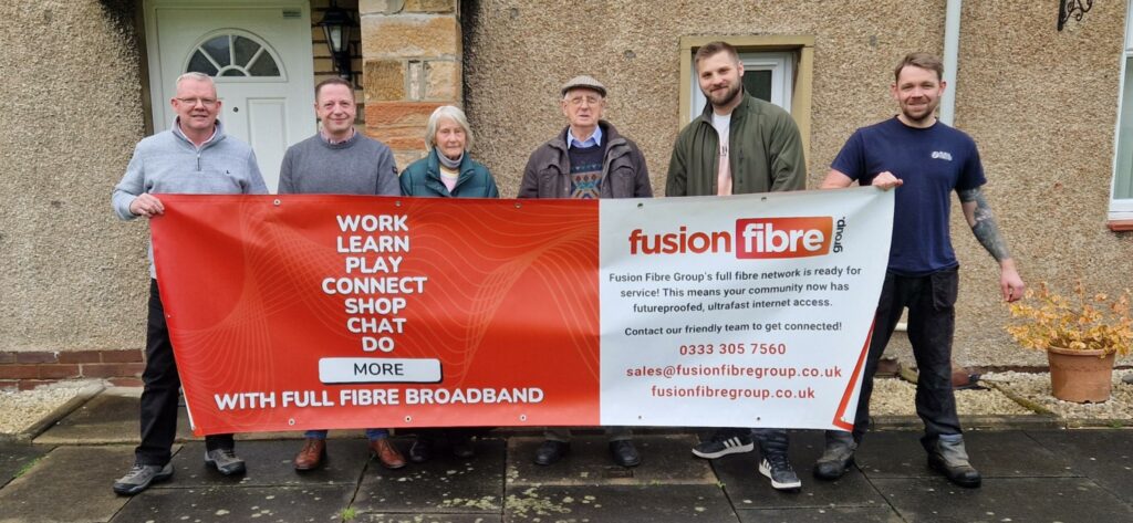 Stocksfield and Mickley's first residents with an ultrafast broadband connection. six people hold up an orange banner infront of a brown house.
